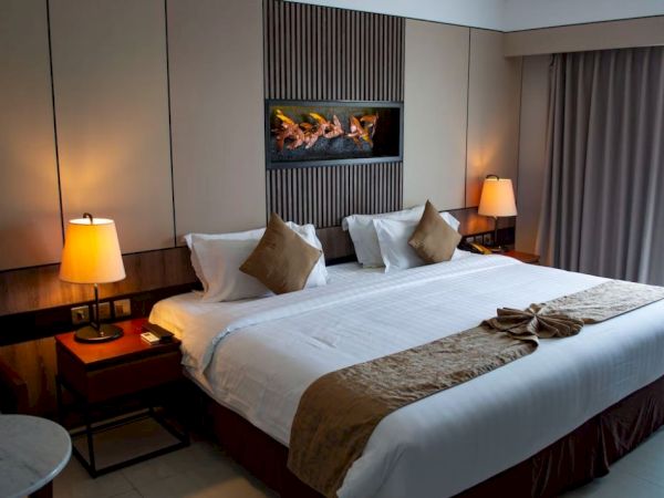 The image shows a well-lit hotel room with a large bed, decorative pillows, and bedside lamps, creating a cozy and inviting atmosphere.
