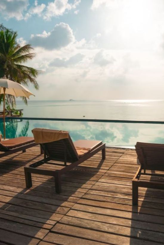 A wooden deck with lounge chairs and umbrellas overlooks a serene infinity pool and ocean, under a partly cloudy sky.
