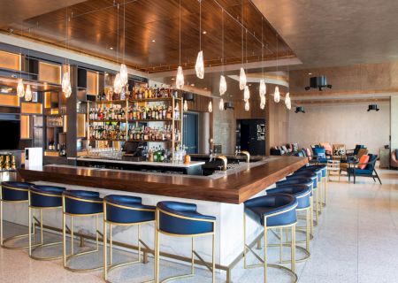 The image shows a modern bar with a wooden counter, bar stools, hanging lights, and stocked shelves, in a spacious, stylish interior.