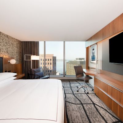 The image shows a modern hotel room with a large bed, a TV, a desk, a chair, and a window with a city view.