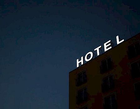 The image shows the exterior of a building at dusk with an illuminated "HOTEL" sign on the rooftop against a dark sky.