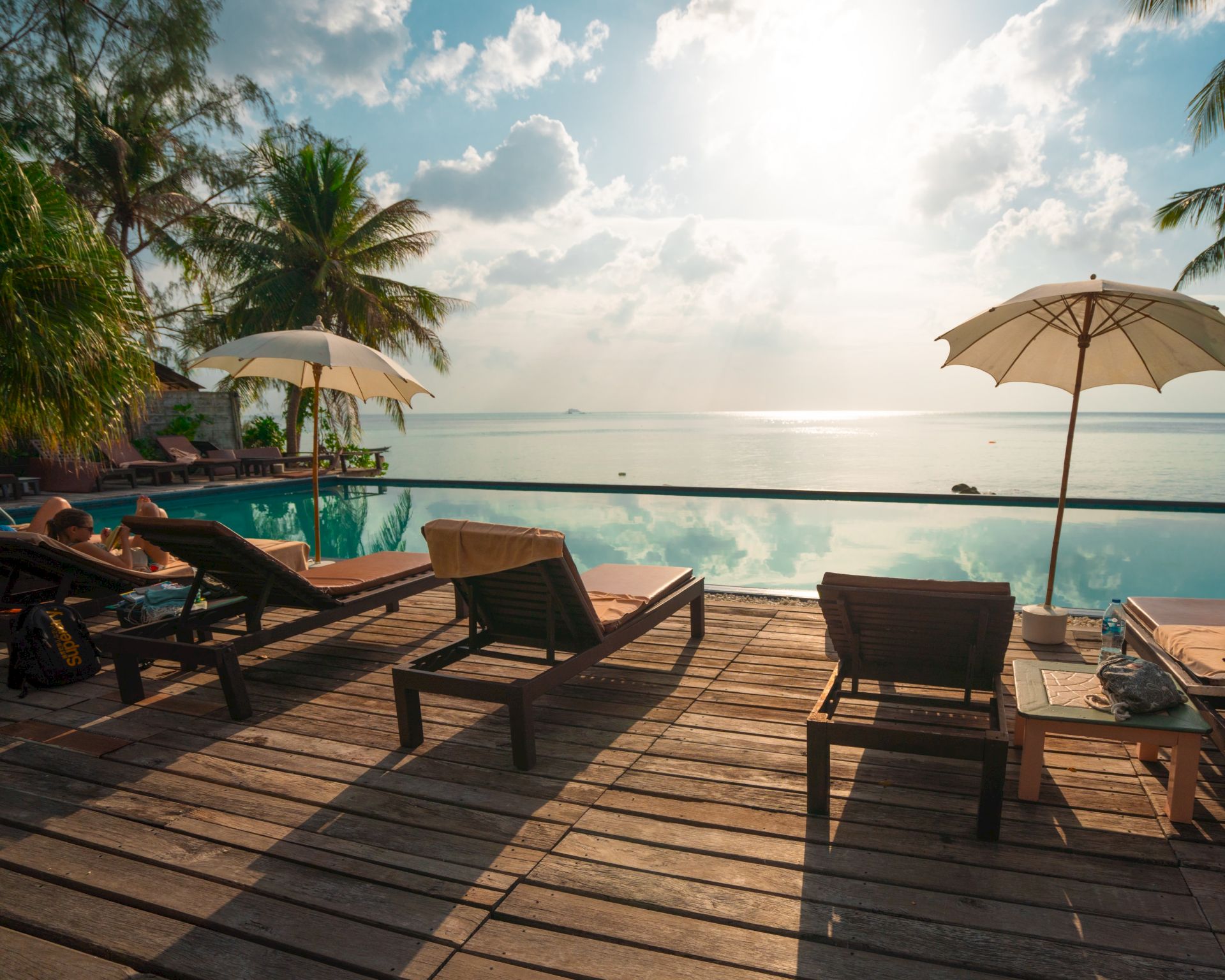 A serene poolside setting with lounge chairs, umbrellas, palm trees, and a view of the ocean under a sunny sky.