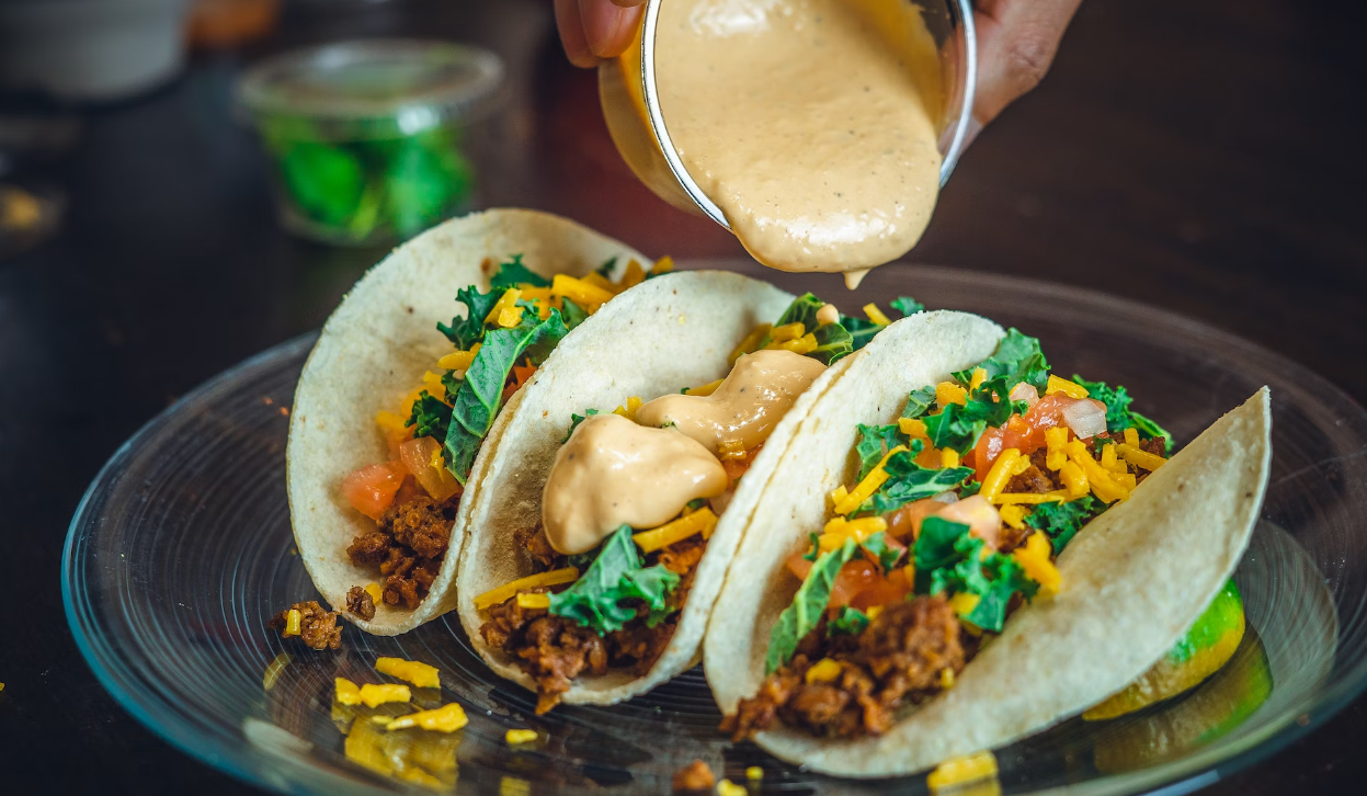 Three tacos filled with meat, cheese, lettuce, and tomatoes on a plate. A hand pours sauce over them from a small container.