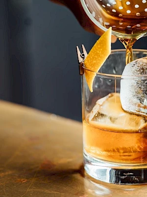 A cocktail is being poured into a glass with ice and a twist of orange peel.