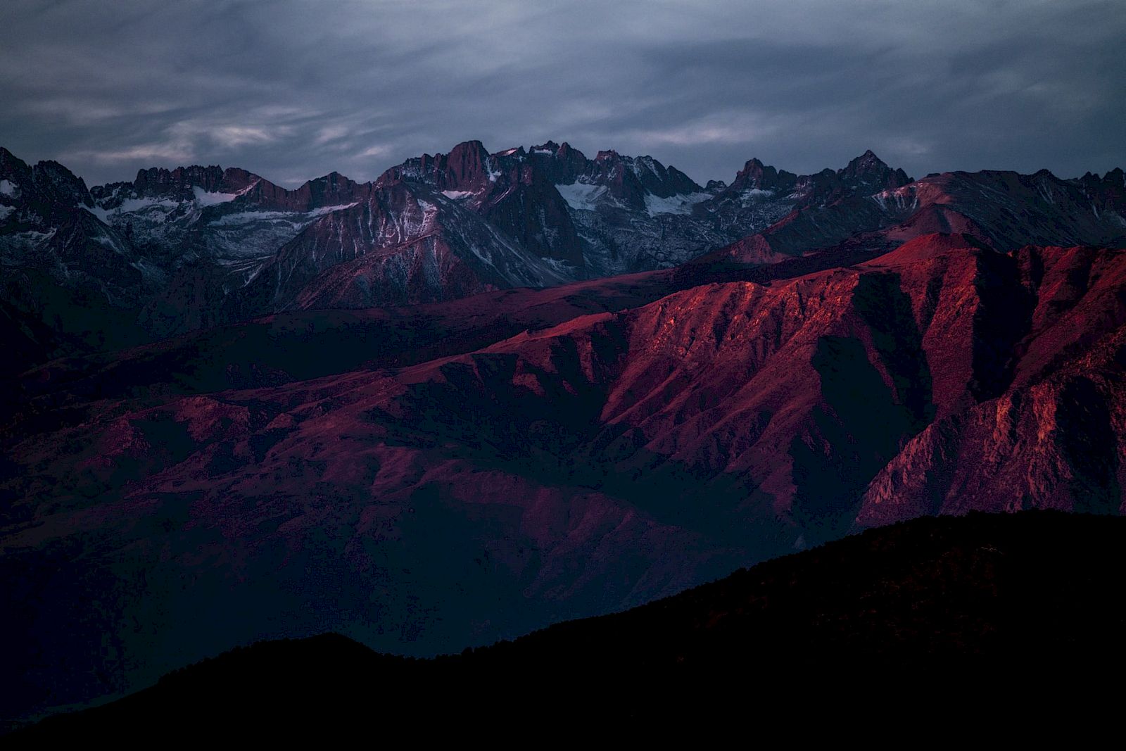 A mountain range under a dramatic sky is illuminated in deep red hues during twilight, creating a striking, moody scene.
