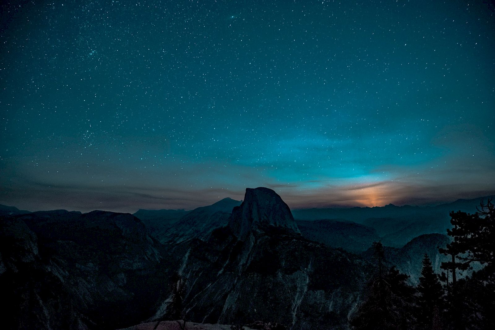 A starry night sky over a mountainous landscape with a prominent peak silhouetted against a dimly lit horizon.