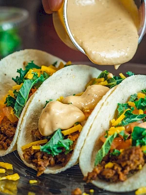 Three tacos with ground meat, cheese, greens, and sauce being poured over them on a plate.