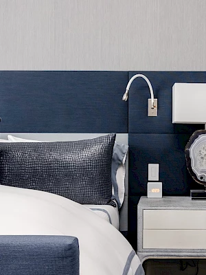 A modern bedroom with a navy headboard, white bedding, and a nightstand featuring a lamp, books, and a decorative item.