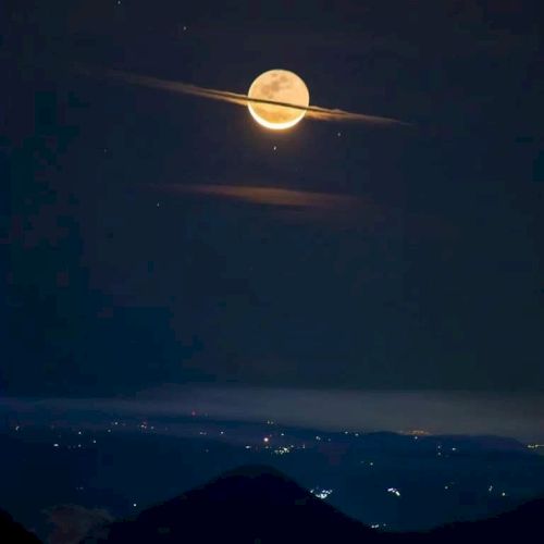 A bright full moon is partially covered by horizontal clouds, resembling Saturn, above a silhouetted landscape with city lights below.