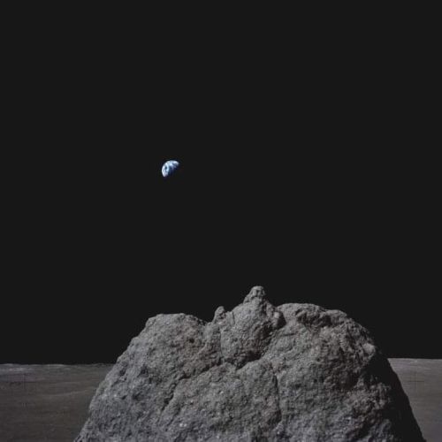 A rock on a barren surface is in the foreground with a small Earth visible against a dark sky in the background.