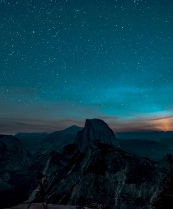 A starry night sky over a mountainous landscape with a prominent peak, possibly during a moonrise or sunset.