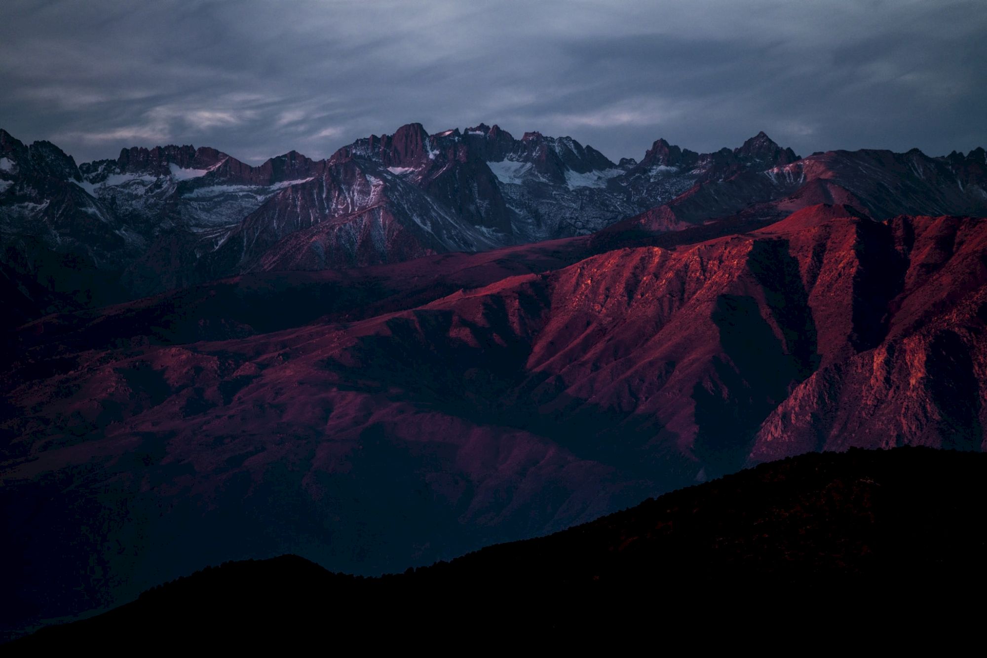 A mountain range is bathed in deep red light, under a cloudy sky, creating a dramatic and moody landscape scene.