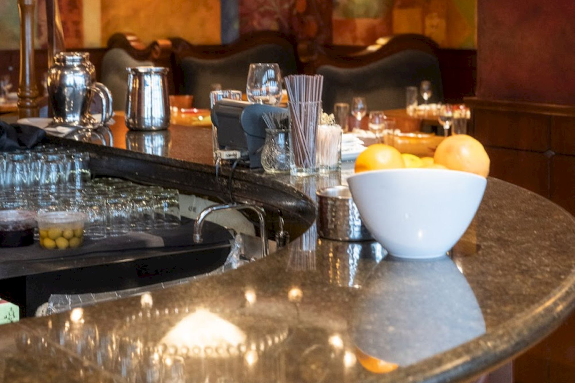 A bar counter with a bowl of citrus fruits, glasses, a pitcher, and various bar tools and accessories displayed neatly.