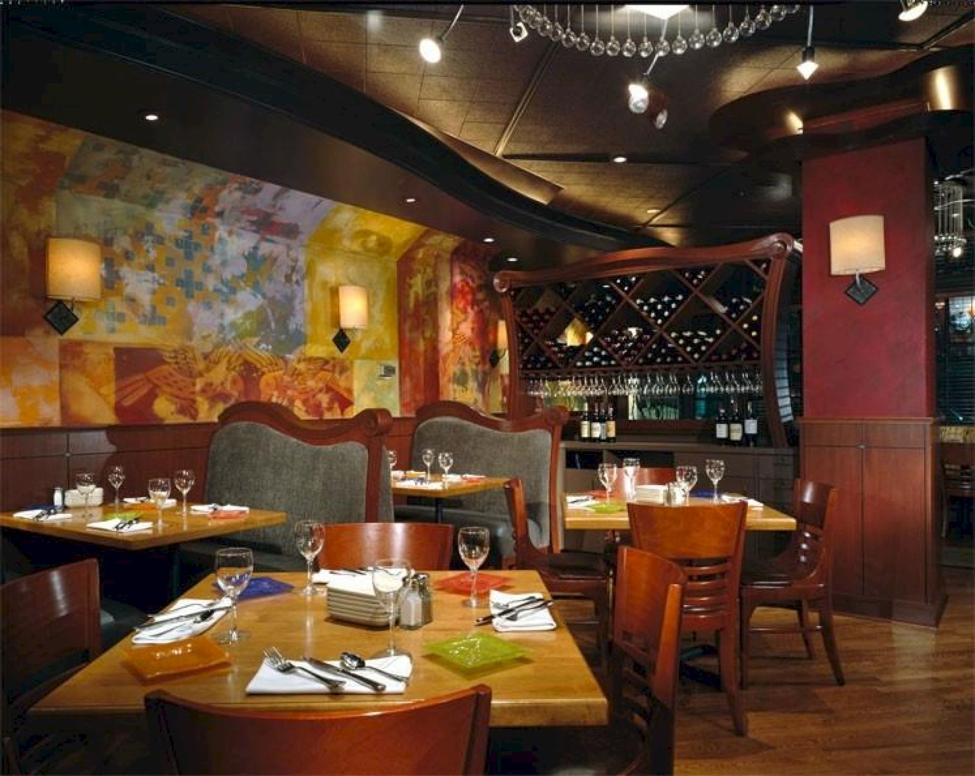 The image shows a restaurant with wooden tables and chairs, colorful wall art, soft lighting, and wine bottles displayed in the background.