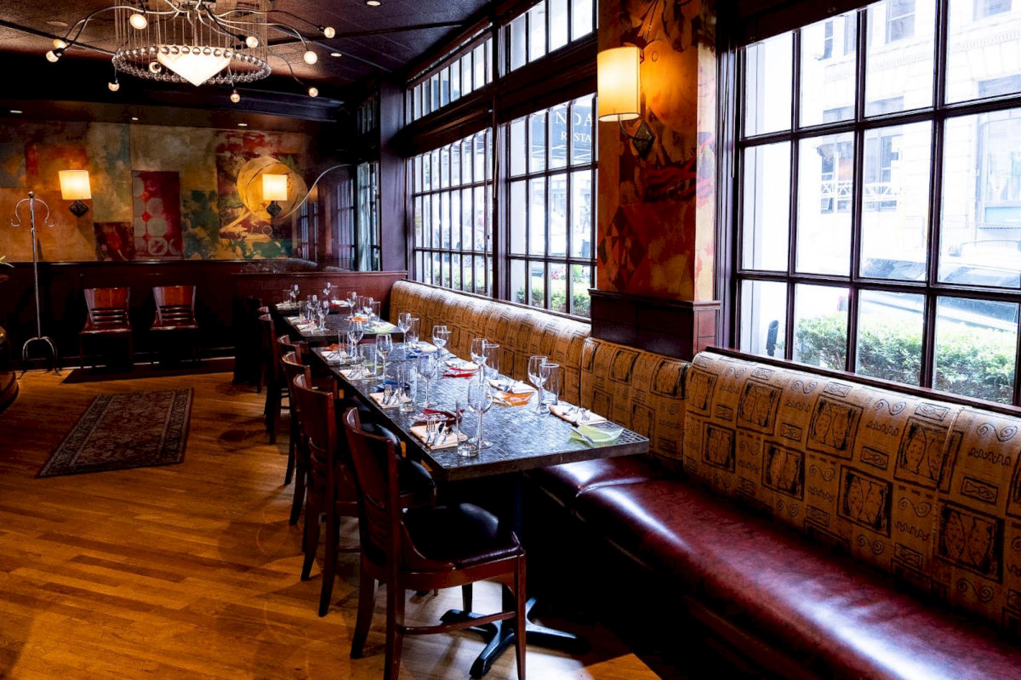 A cozy, elegant restaurant interior with a long dining table, set for multiple guests, large windows, and warm lighting.