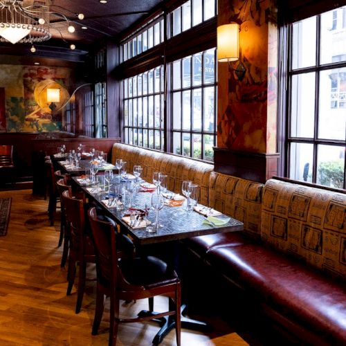 The image shows a restaurant interior with a long table set for dining, featuring upholstered seating and large windows along one wall.