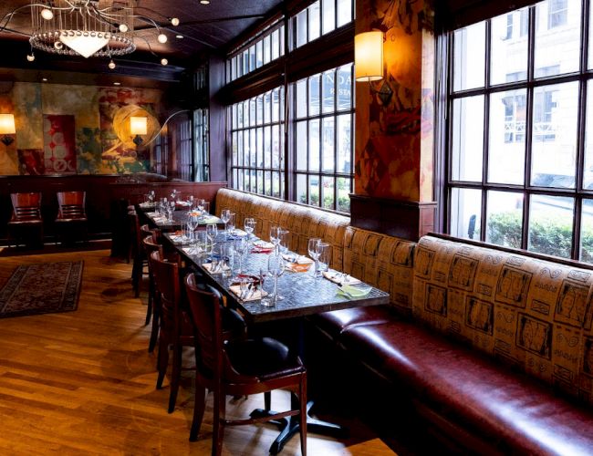 The image shows a restaurant interior with a long table set for dining, featuring upholstered seating and large windows along one wall.