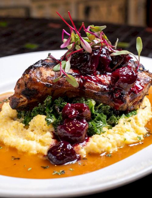 Grilled meat on mashed potatoes with greens, topped with berry sauce and microgreens, served on a white plate with a light sauce base.
