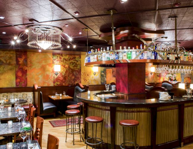 A cozy bar/restaurant with a curved wooden bar, hanging shelves of bottles, warm lighting, and colorful mural walls, plus dining tables nearby.
