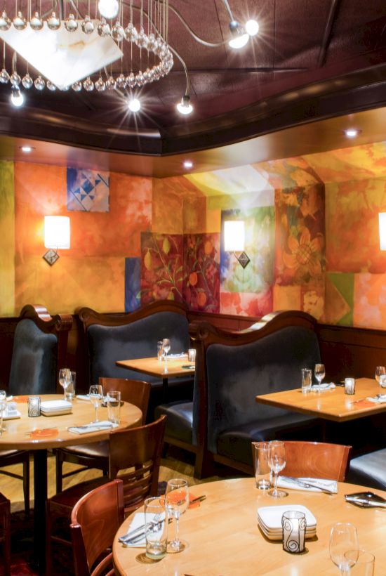A stylish, colorful dining room with round wooden tables, plush black booths, warm lighting, and vibrant mural walls&mdash;cozy and upscale.