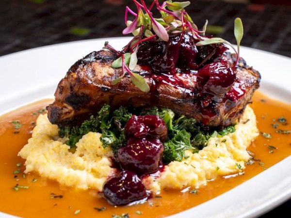 Grilled meat topped with berry sauce on mashed potatoes and greens, garnished with microgreens, served on a white plate.