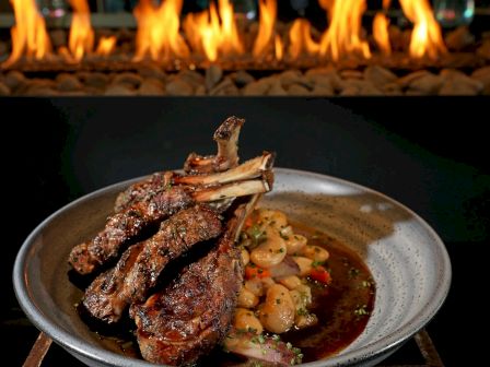 A dish with grilled lamb chops served on a plate, accompanied by a vegetable medley, set against a backdrop of a burning fire.