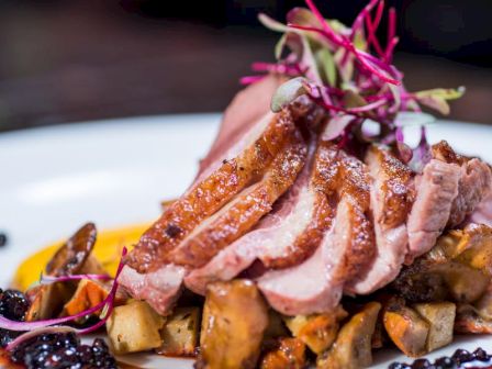 A gourmet dish featuring sliced duck breast on a bed of mushrooms, garnished with microgreens and a berry sauce, presented on a white plate.