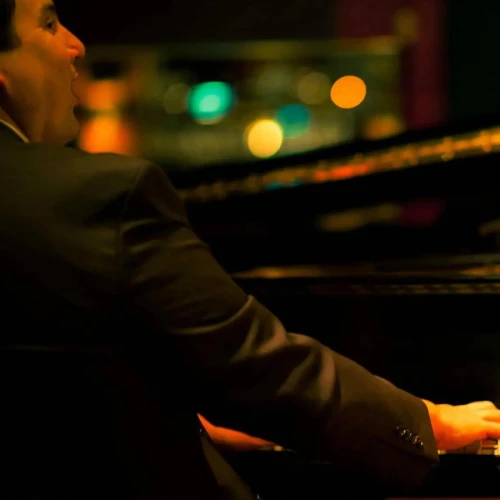 A person in a suit is playing a piano, with colorful lights in the background creating a warm ambiance.