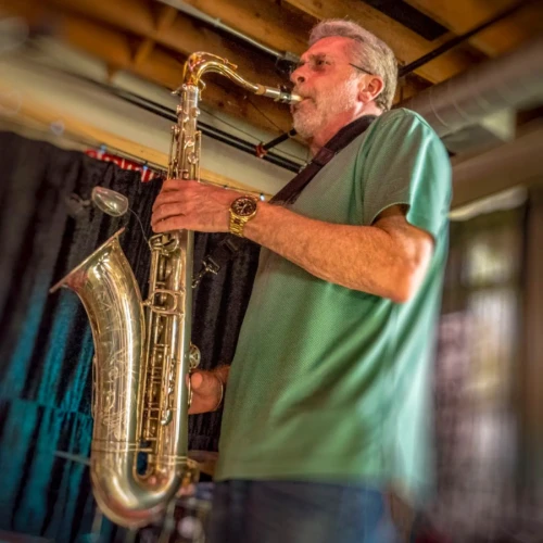 A person playing a saxophone indoors, wearing a green shirt and glasses, with a blurred background and exposed ceiling beams.