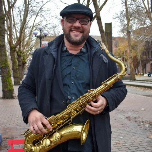 A person is holding a saxophone while standing outdoors on a tree-lined sidewalk, wearing glasses, a hat, and a coat.
