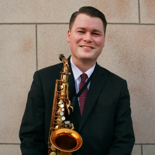 A person in a suit is holding a saxophone and smiling in front of a tiled wall.