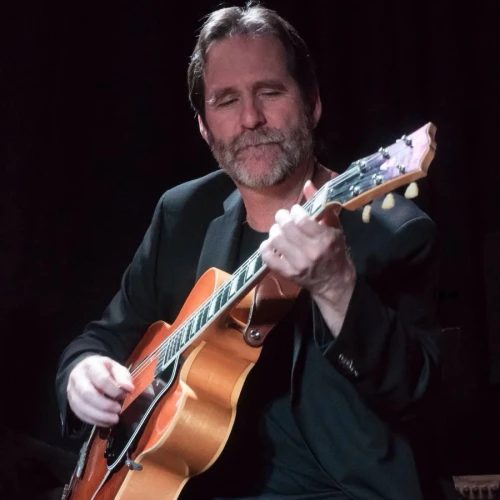 A person is playing an acoustic guitar while seated, wearing a dark blazer, against a dark background.