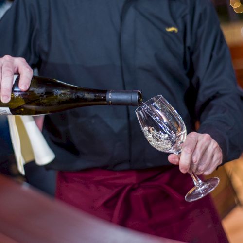 A waiter pours wine from a bottle into a glass at a bar, wearing a dark shirt and red apron, in a fine-dining setting.