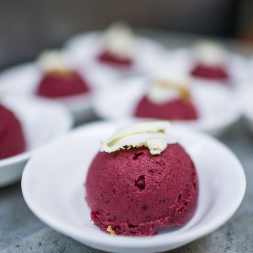 A scoop of deep red sorbet topped with a thin white garnish sits in a white dish, with several similar scoops blurred in the background.