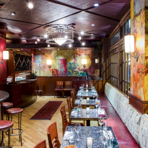 A stylish, cozy restaurant interior with a long row of tables set for dining, warm lighting, artwork on the walls, and a bar area to the left.