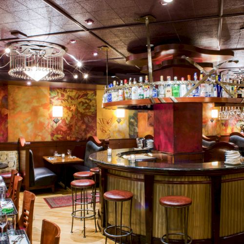 A stylish, cozy bar with a curved wooden counter, hanging lights, and colorful art walls; bar stools line the counter and tables sit nearby.