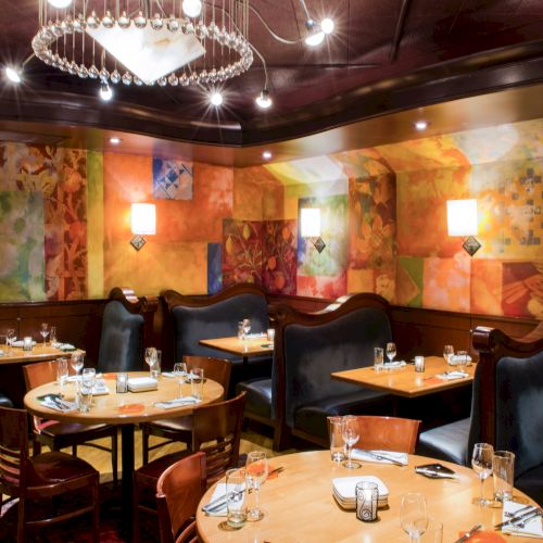 A cozy upscale dining room with round wooden tables, black booth seating, warm colored murals, soft lighting, and a chandelier overhead.
