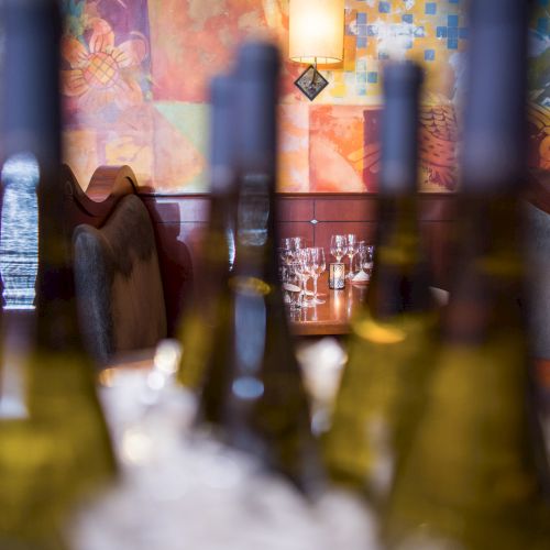 A dimly lit restaurant with blurred wine bottles in the foreground, cozy booths, and colorful wall art in the background, tables set for dinner.
