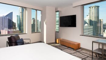 A modern hotel room with large windows, city views, a bed, chair, and a wall-mounted TV.