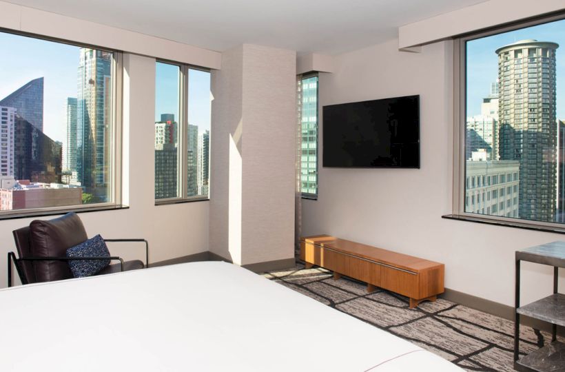 A modern hotel room with large windows, city views, a bed, chair, and a wall-mounted TV.