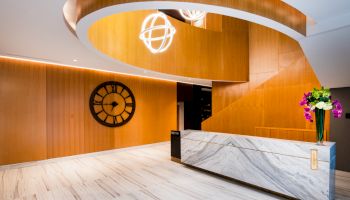 A modern lobby with a marble reception desk, wood paneling, clock on the wall, spiral ceiling, and bright ceiling lights.