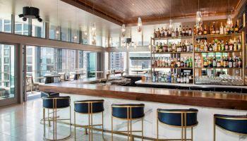 A modern bar with stylish seating, well-stocked shelves, and large windows overlooking a cityscape.