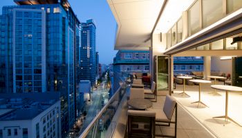 A modern balcony at dusk overlooks a city street flanked by tall buildings, featuring outdoor seating and tables.