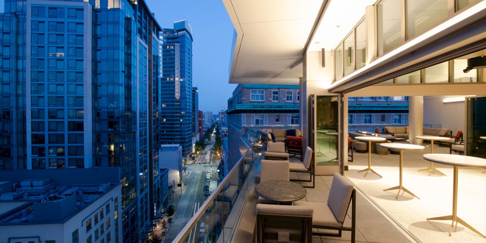 Rooftop terrace with modern seating, overlooking a city skyline at dusk. Illuminated interior visible through large glass windows.