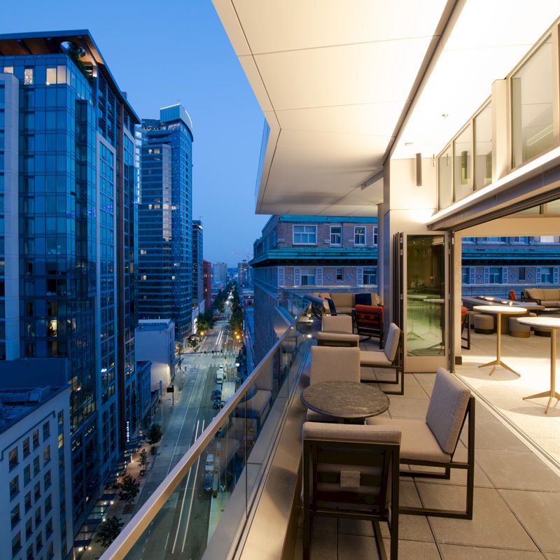 The image shows a modern rooftop terrace with seating, overlooking a cityscape with tall buildings at dusk.