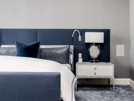 A modern bedroom features a blue headboard, white bedding, a nightstand with a lamp and framed artwork, and a plush carpet.