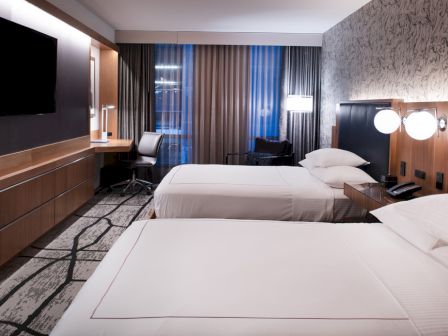 A modern hotel room with two beds, a wall-mounted TV, desk area, and stylish decor featuring patterned carpet and lighting fixtures.