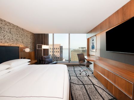 A modern hotel room with a large bed, desk, flat-screen TV, and city view through a window.