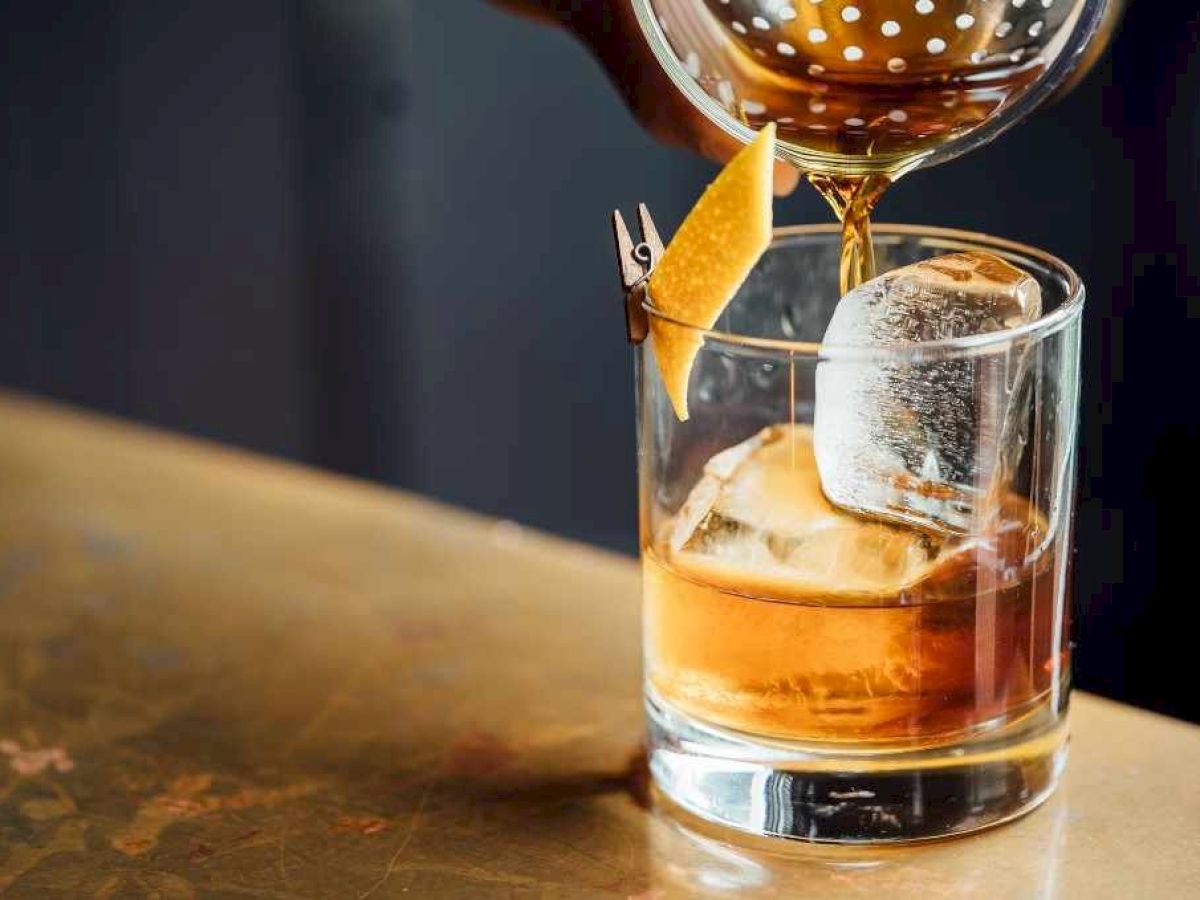 A drink being poured into a glass with ice and a citrus peel on a bar counter.