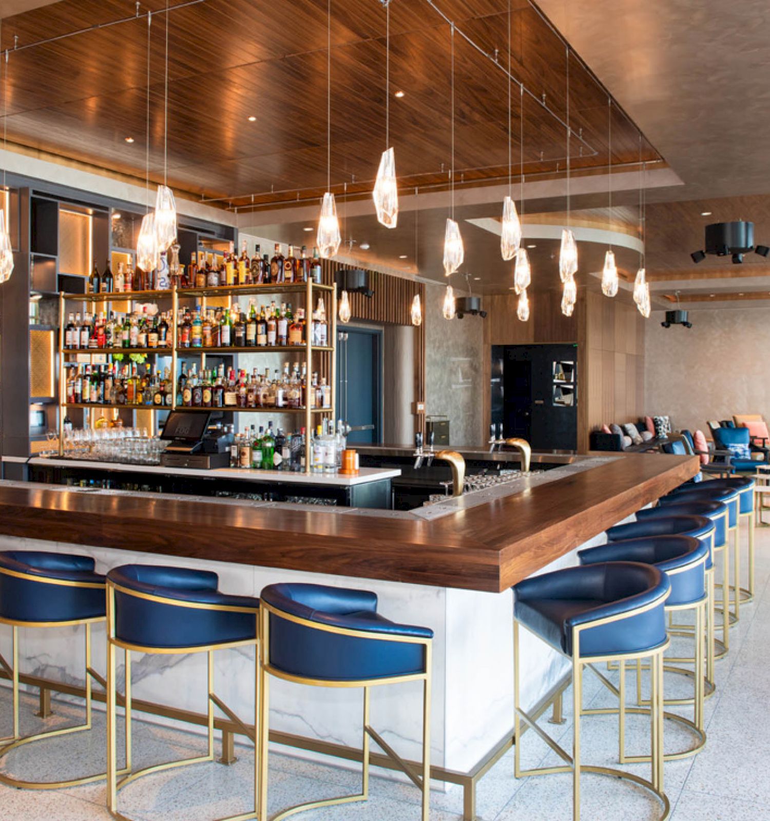 The image shows a modern bar with high stools, a central counter stocked with bottles, and stylish pendant lighting in a spacious setting.